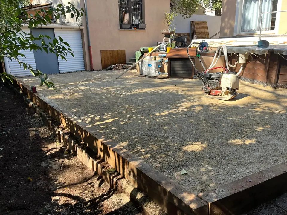 Plateforme en gravier compacté bordée de traverses en bois, plaque vibrante posée, devant une maison