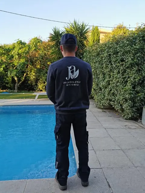 Paysagiste de dos, en sweat brodé au nom de l’entreprise, devant une piscine bordée d’une haie