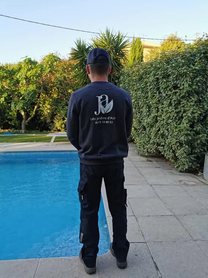 Paysagiste de dos, en sweat brodé au nom de l’entreprise, devant une piscine bordée d’une haie