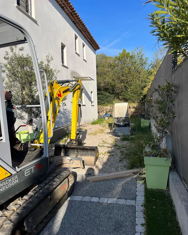 Mini-pelle jaune engagée dans le passage étroit entre une maison et un mur, avant l’aménagement