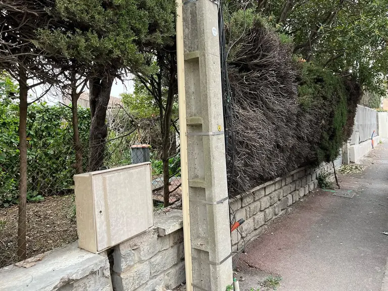 Muret en pierre bordant une rue, poteau de clôture et coffret encastré, haie derrière