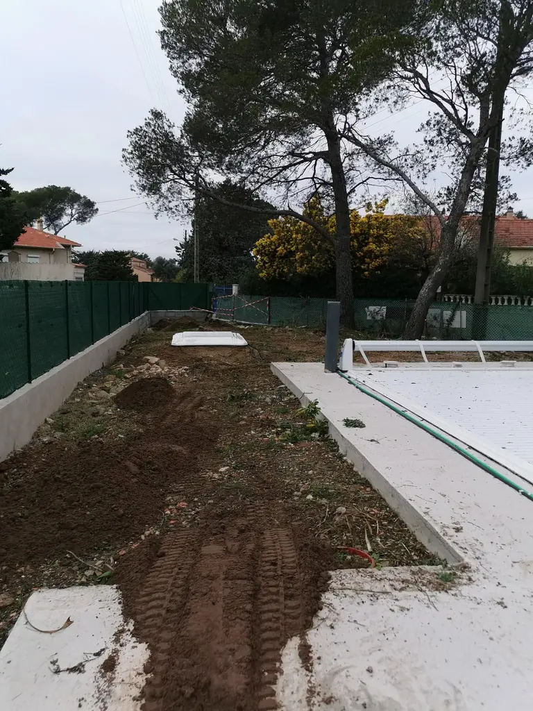 Terre remuée le long d’une piscine bâchée, avant la pose de la pelouse, clôture verte en fond