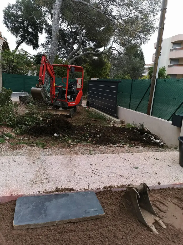 Mini-pelle rouge au travail dans un petit jardin, terre retournée, clôture verte en fond